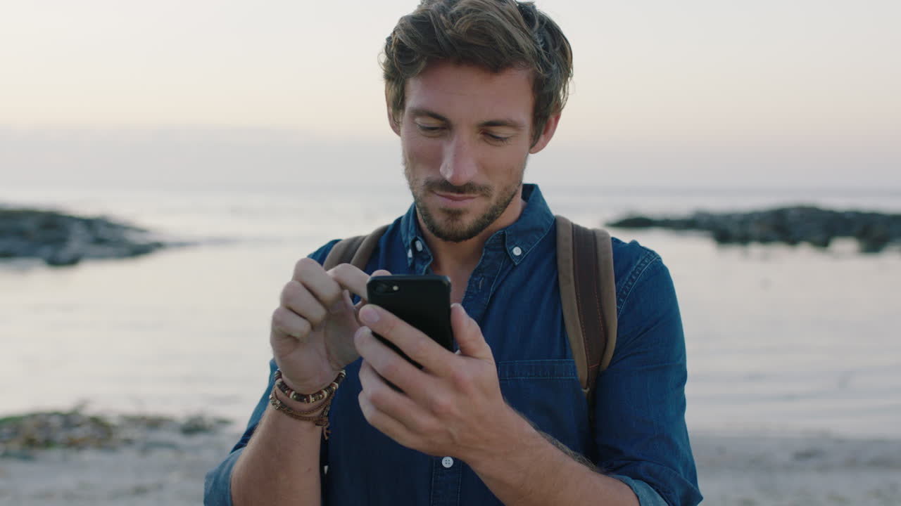 ritratto di un attraente uomo caucasico che usa uno smartphone per scrivere messaggi sulla spiaggia
