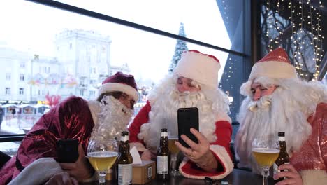 Three Santa Claus Looks at the Smartphone Screen with his Eyes Wide Open three Santa Claus Looks at the Smartphone Screen with his Eyes Wide Open Men in Santa Claus Costume Resting in a Cafe Christmas Time