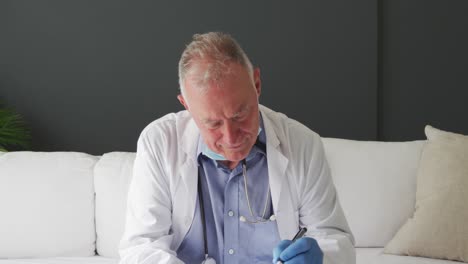 médico de sexo masculino caucásico con la máscara bajada tomando notas mientras hace una videollamada