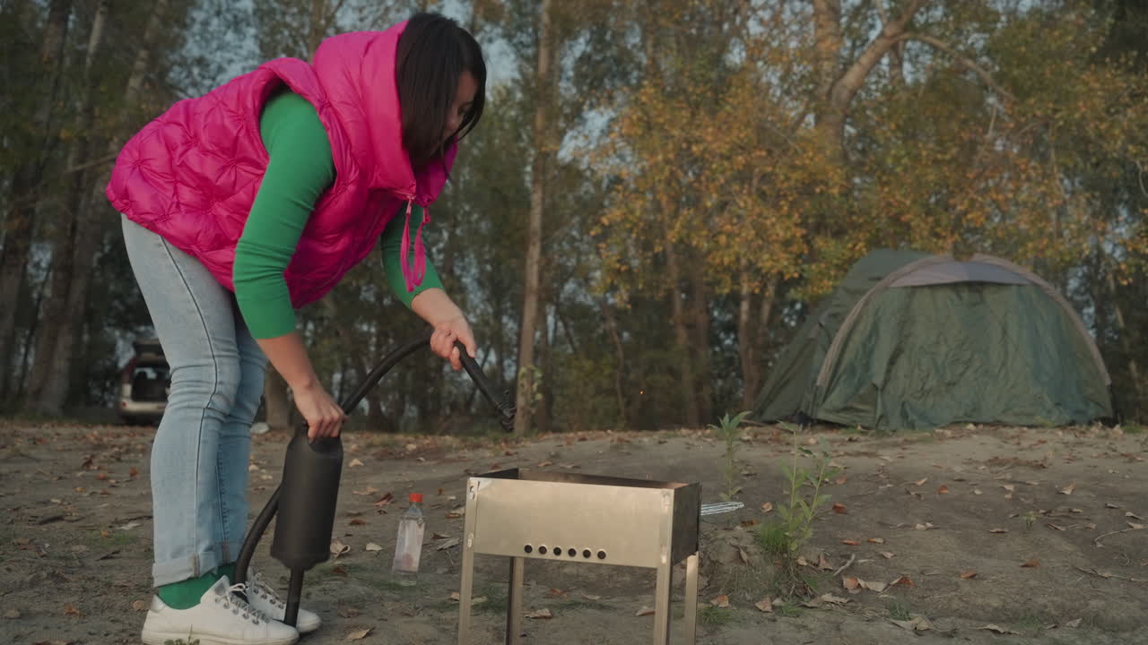 Kvinne forbereder bærbar ovn ved siden av teltet, campingscene i høstskog med grønt kuppeltelt i bakgrunnen, camper i rosa vest og jeans heller brensel nær en kompakt grill, arrangerer grillkull.