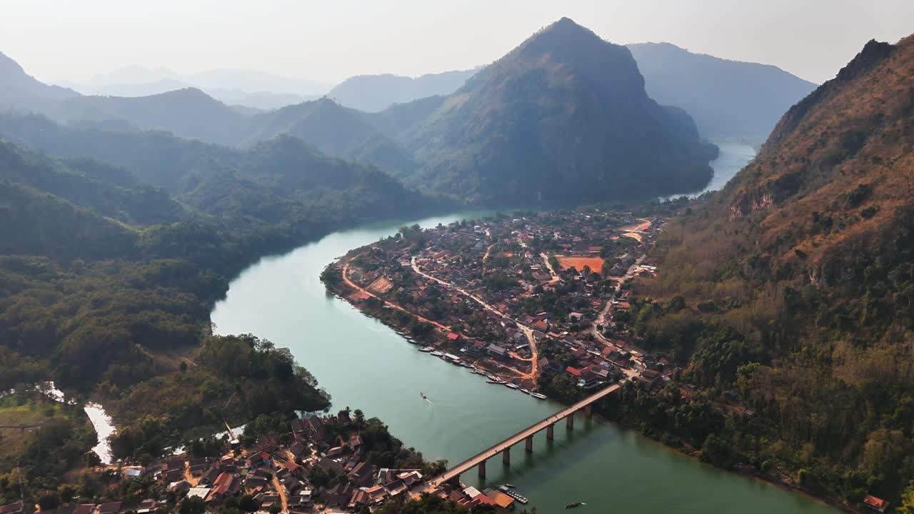 Aerial view of Nong Khiaw, Laos, with a winding river encircling the village, mountains rising around the valley, and a bridge connecting the riversides under clear daylight in a tranquil setting