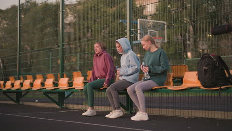 group of ladies sitting on stadium seats with water bottles, two crossing legs while one stretches her leg forward, casual outdoor activity, fitness, and relaxation scene