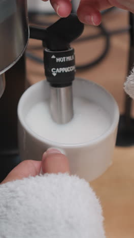 mujer azota leche para capuchino en la máquina de café. dama en bata de baño azota la leche en espuma usando capuchinador de cafetera. proceso de fabricación de espuma de leche