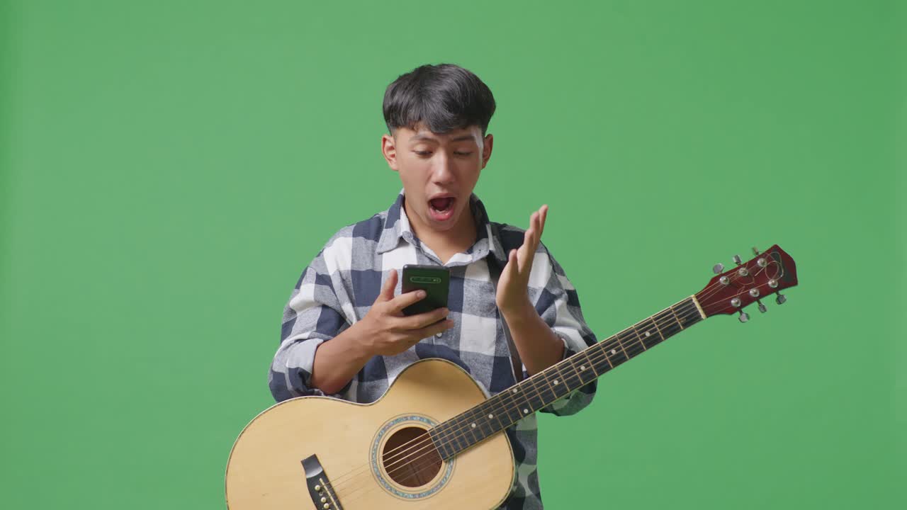 스마트폰을 사용하고 그린 스크린 배경에 서서 와우라고 말하는 기타를 가진 아시아 십대