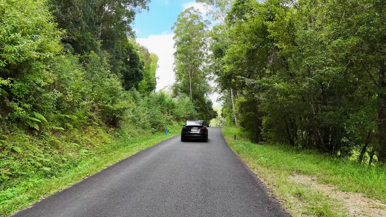 A car travels along a serene, tree-lined road in Bellingen, NSW. Bright daylight illuminates the lush greenery and winding path
