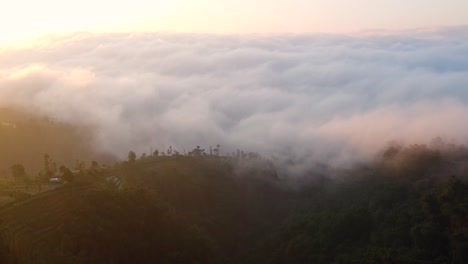 Vista-Aérea-De-La-Montaña-Cubierta-Y-El-Denso-Paisaje-Nuboso-Que-Oculta-El-Valle-En-Indonesia-Durante-El-Amanecer-Dorado