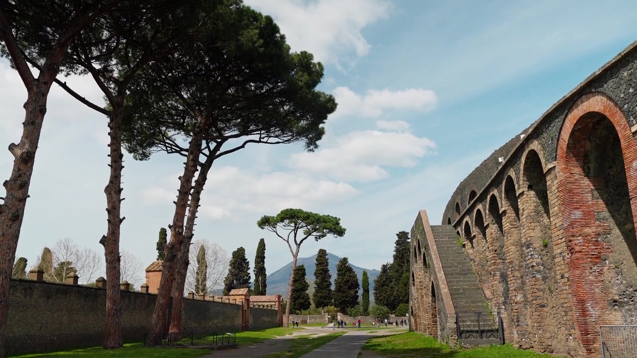Arcata esterna dell'Anfiteatro di Pompei vicino alla Palestra Grande, con archi in mattoni, scala in pietra, pini marittimi, cipressi e il Vesuvio in lontananza.