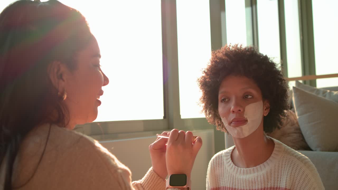 Hispanic mom brushing mask onto daughter's cheeks for pampering in living room, copy space
