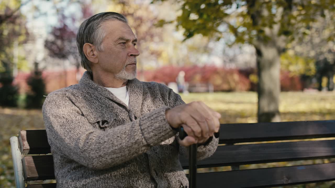hombre caucásico viejo serio sentado en un banco en el parque durante el otoño