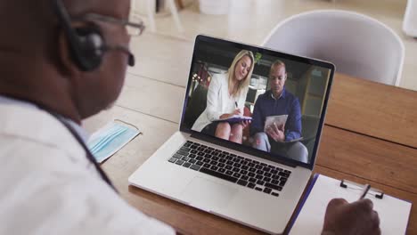 African-american-male-doctor-wearing-phone-headset-taking-notes-while-having-a-video-call-on-laptop