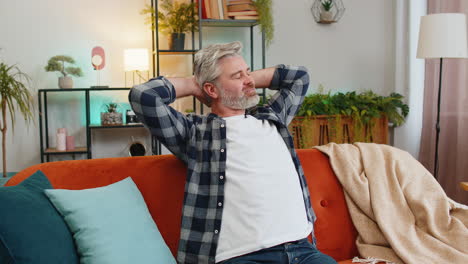 tired senior middle-aged man stretching arms and resting relaxing on sofa in living room at home