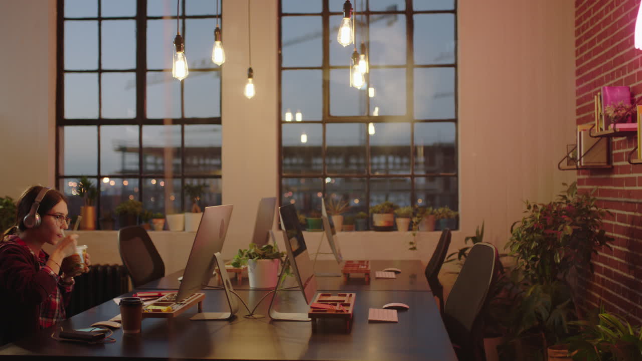 젊은 백인 사업가 여성 학생 컴퓨터를 사용하여 늦게 일하는 타이핑 연구 프로젝트 온라인 브라우징 브레인스토밍 기한 트렌디한 사무실에서 밤에 헤드폰을 착용