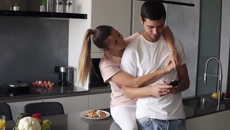 A-man-focused-on-his-phone-while-his-girlfriend-wife-is-trying-to-get-his-attention,-hugs-him-from-the-back.-A-young-couple-standing-embraced-in-casual-clothes-on-the-modern-kitchen