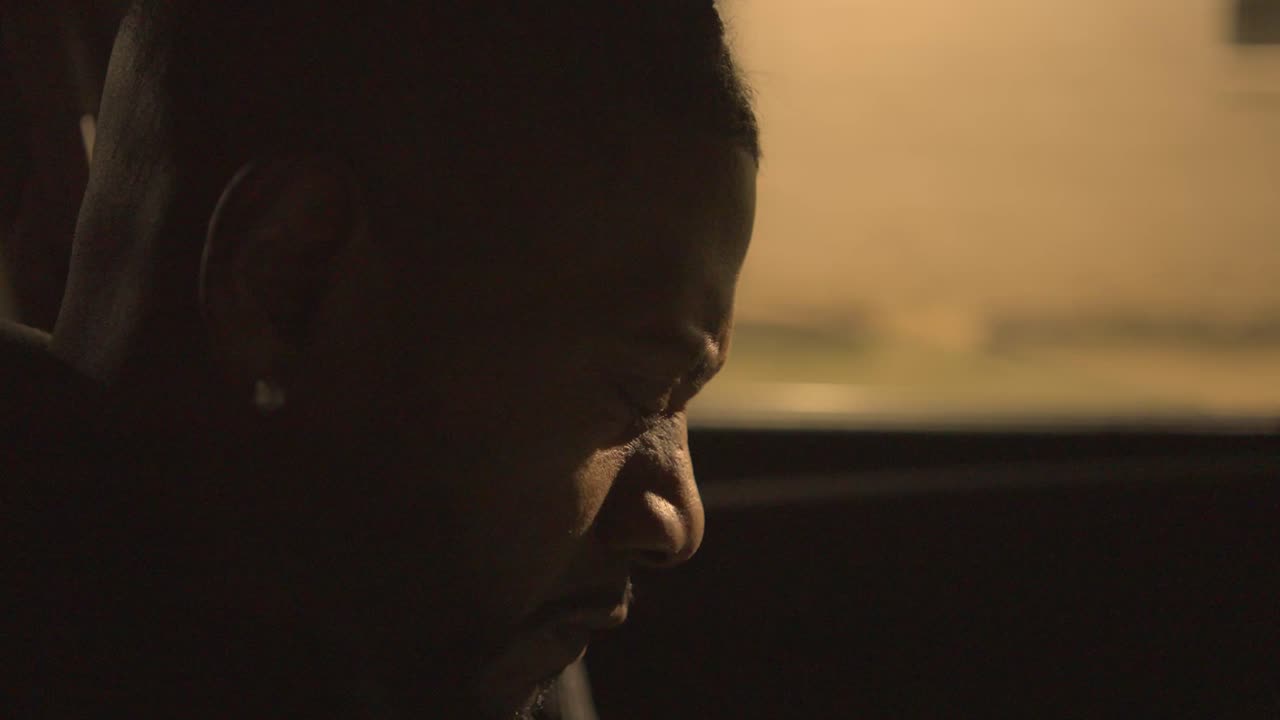 um homem negro sentado no banco do motorista de um carro à noite balança a cabeça e cobre o rosto em descrença