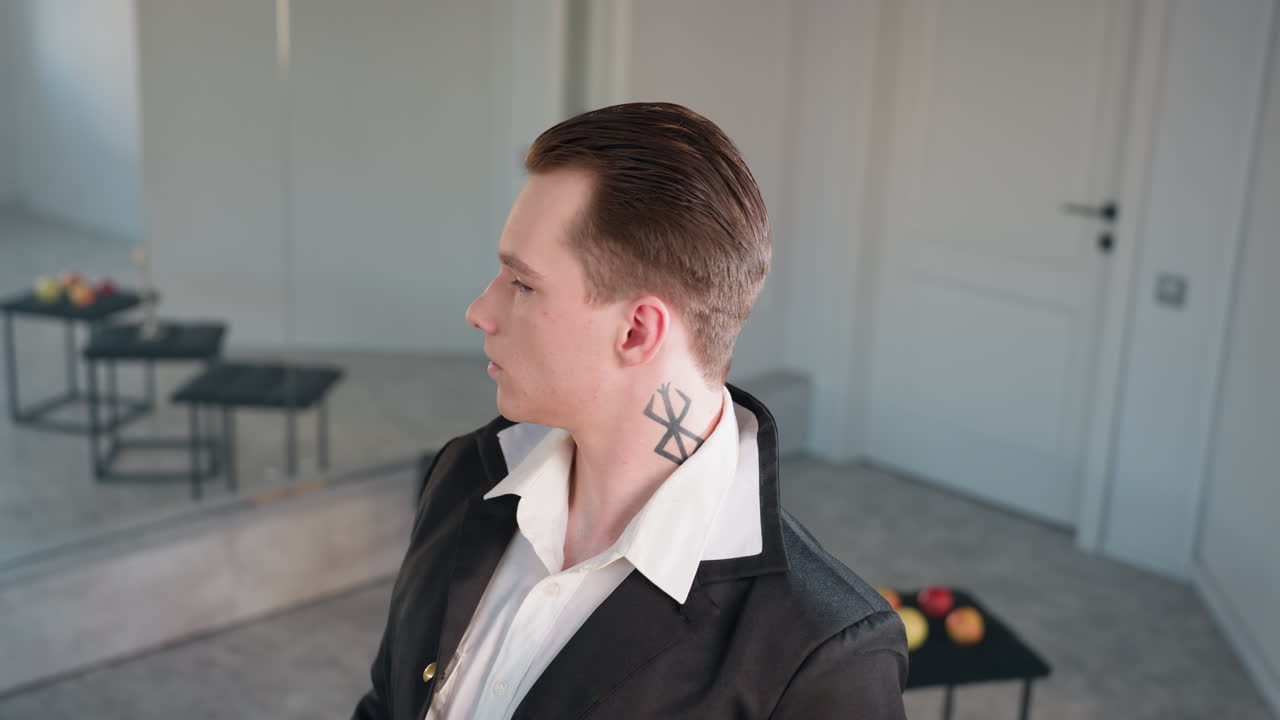 young man with slicked hair and neck tattoo stands confidently in studio, three stools hold apples and candle, large mirror reflects scene, soft light casts elegant mood