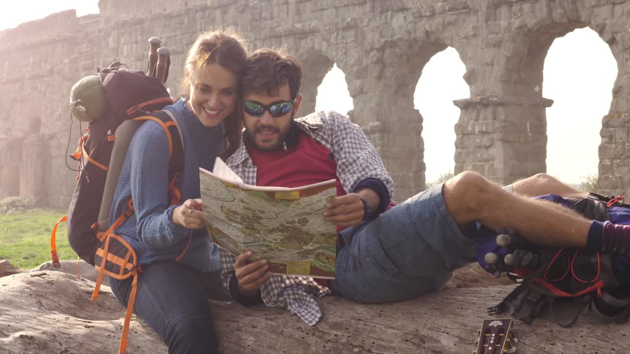pareja feliz de mochileros turistas sentados acostados en un tronco de madera leyendo una guía de mapas apuntando a las antiguas ruinas del acueducto romano en el parco degli aquedotti parque en roma al amanecer romántico con saco de dormir cámara lenta