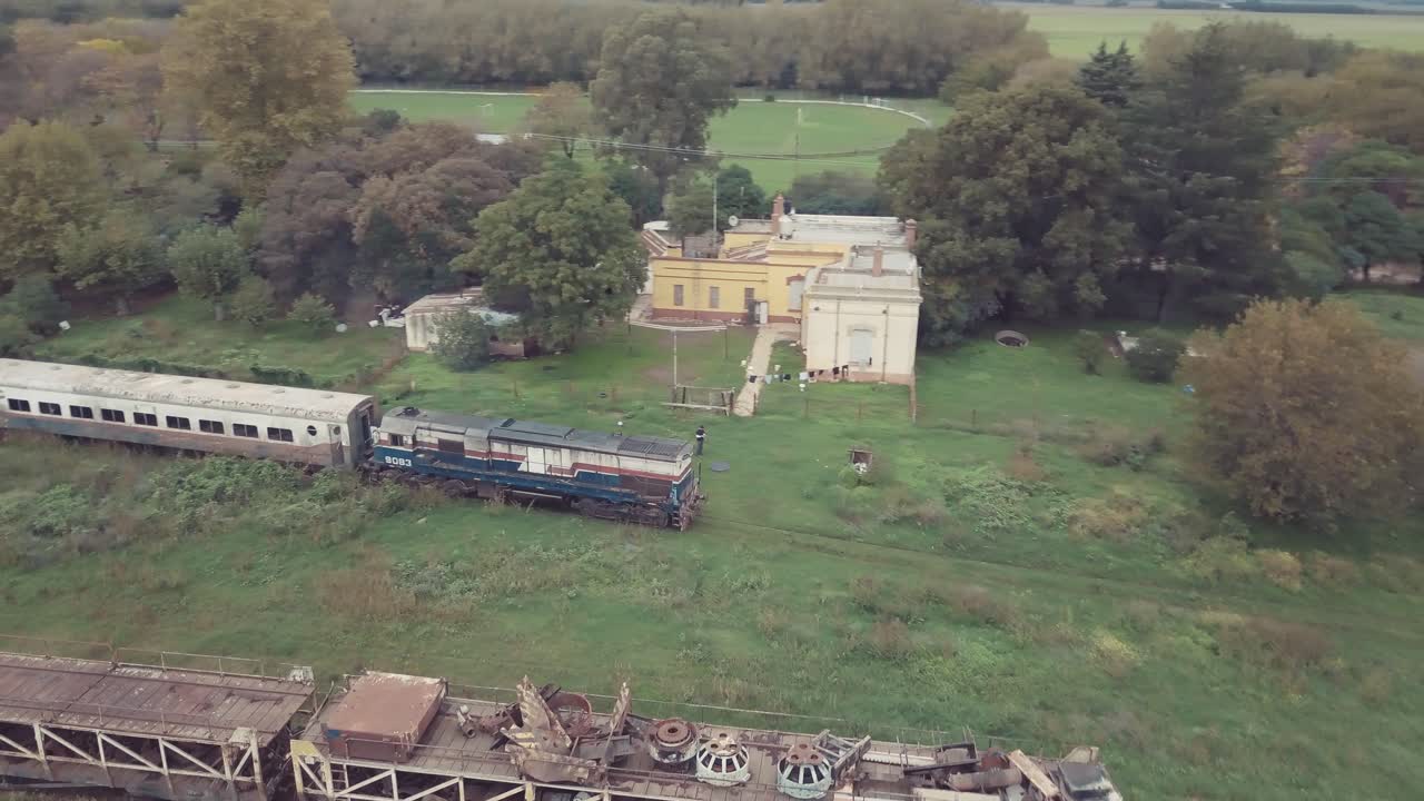 aerial view of an old train in motion with a rural school background and abandoned trains in Buenos Aires province