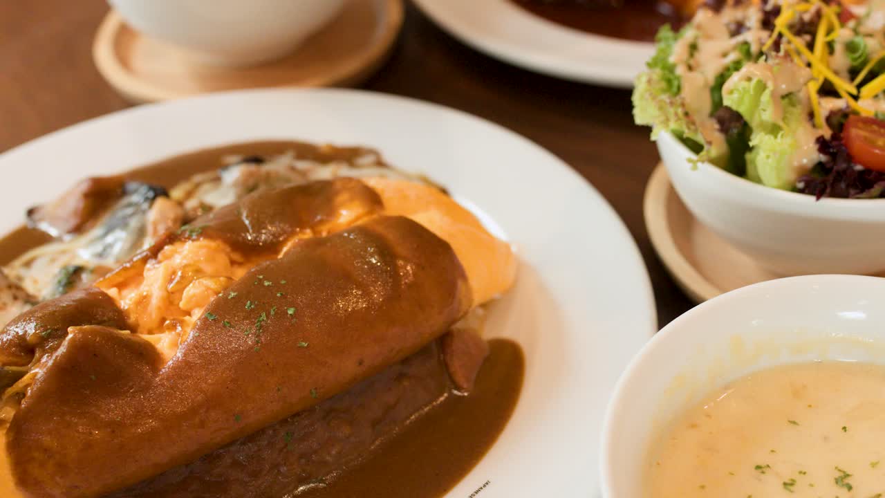Hand slices omelette rice with brown sauce, soup, and salad in bright restaurant setting