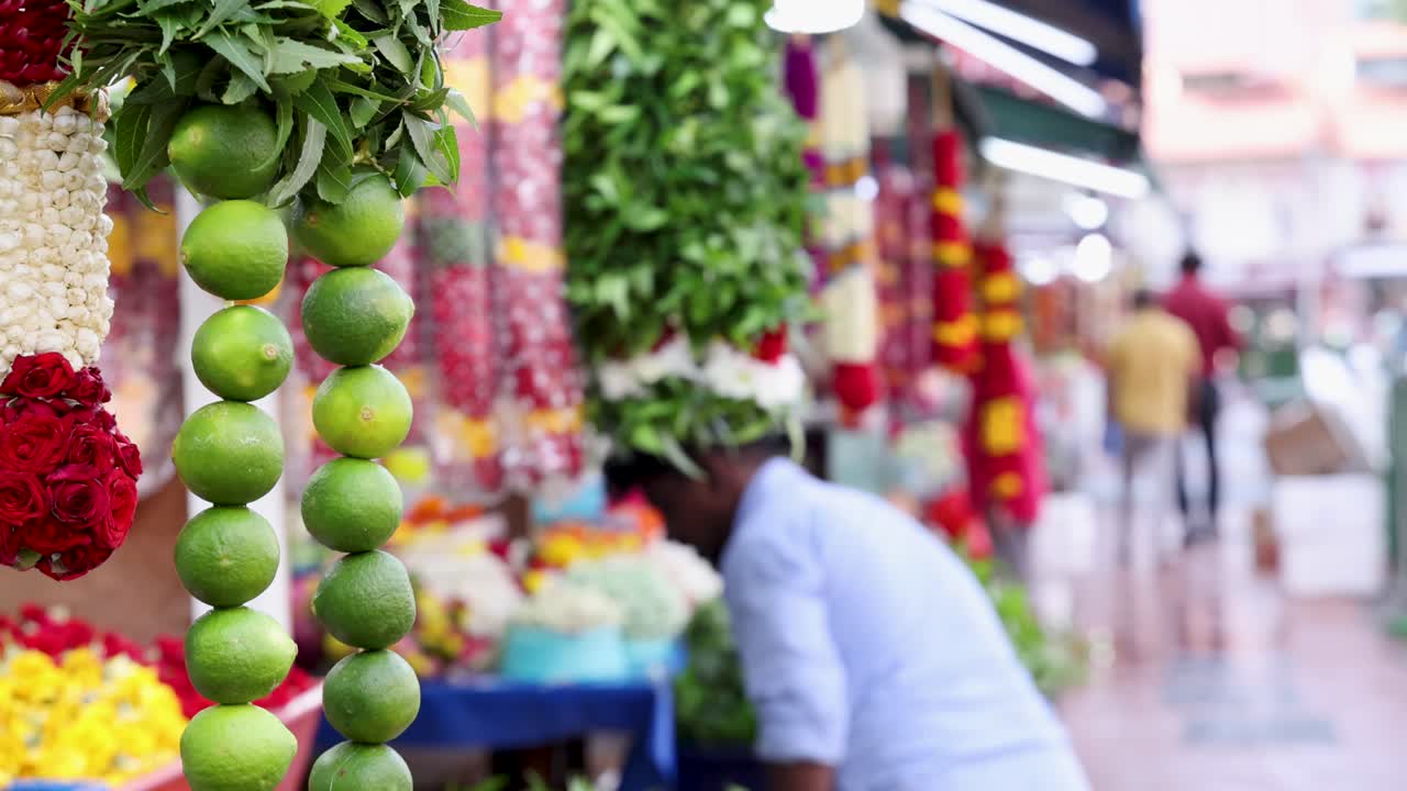 A vendor arranges vibrant flower garlands at an outdoor market stall in Little India, Singapore. Daylight, shallow depth of field, and handheld camera movement create a lively street scene