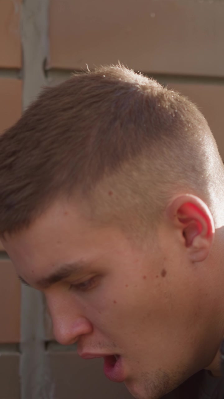 Primo piano di un giovane uomo che esala vapore all'aperto, con capelli corti e texture della pelle visibile. Il soggetto rimane immobile contro un muro esterno, mostrando tratti del viso coerenti, inclusi nei.