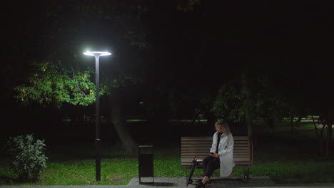 young girl seated alone on park bench at night operating phone beneath glowing streetlamp, surrounded by trees and quiet ambiance, dressed in white coat with umbrella beside her on paved ground