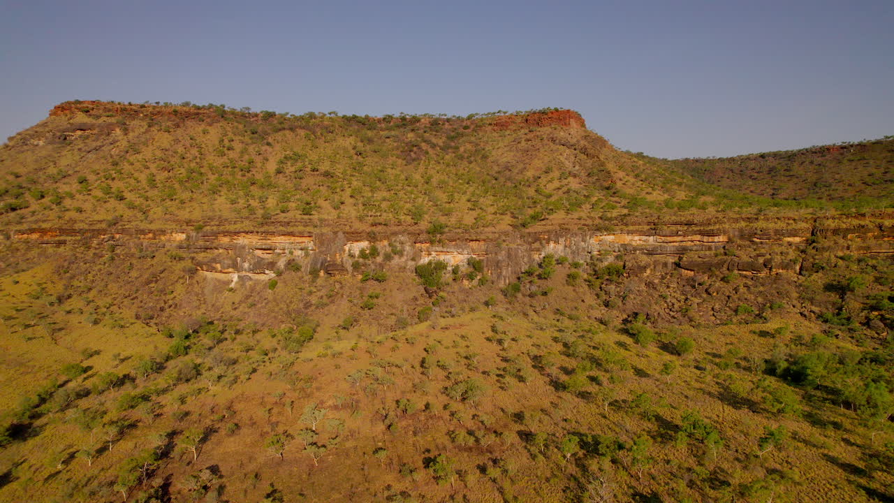 Un drone sorvola il Saddle Creek Campground nella luce del sole serale, rivelando scogliere, boscaglia verde, colline scoscese e la spettacolare catena montuosa nella natura selvaggia del Northern Territory lungo la Victoria Hwy.