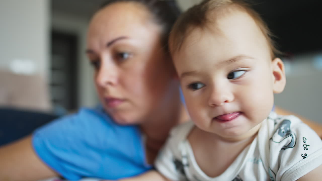 Mother and her baby boy beside. Little cite kid looks into camera with interest showing his tongue. Close up. Blurred backdrop.