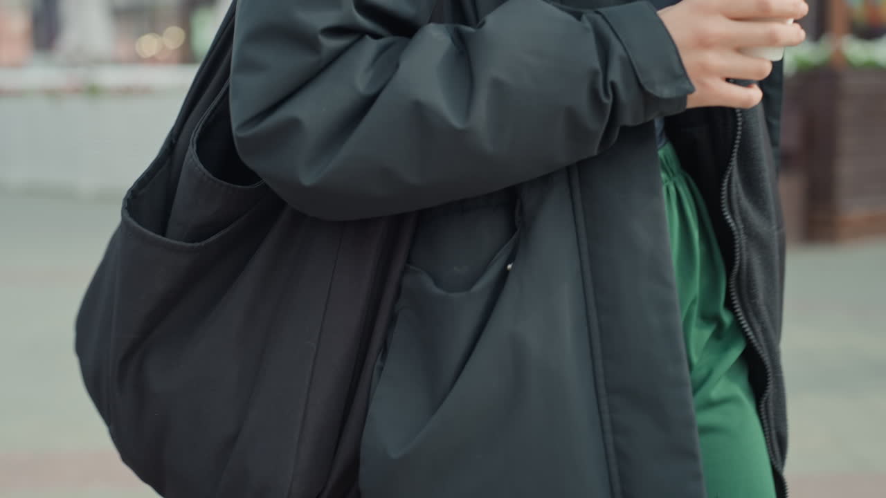 黒いジャケットを着てコーヒーを持ち、肩に大きな黒いトートバッグをかけた人物、クローズアップしたミッドセクション、落ち着いたトーン、都会の歩道、柔らかい昼光、かすかな動き、孤独な雰囲気、カジュアルなストリートウェア。