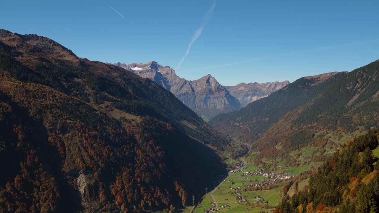 Aerial drone footage showing a calm mountain valley with a small town below. Captured on a clear autumn day, the scene highlights colorful forests and towering peaks under soft sunlight