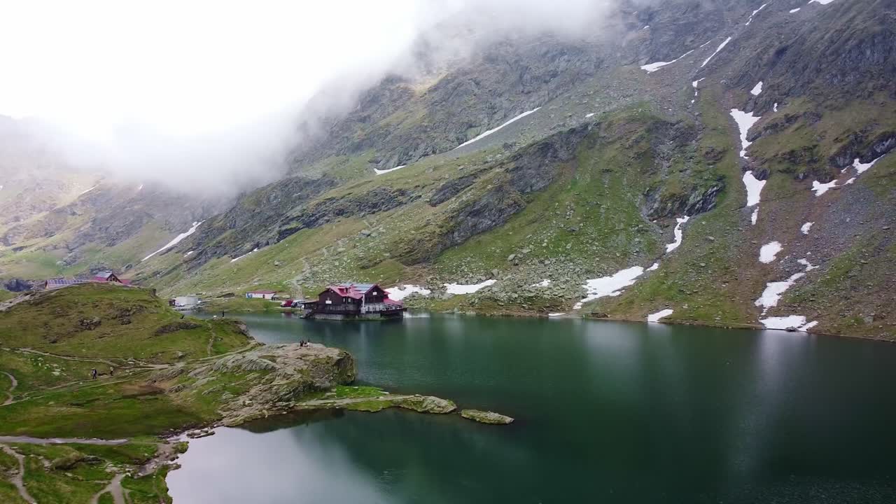 Drone aerial view over Transfăgărășan, winding road, Balea lake lodges, rocky cliffs and mountain mist.