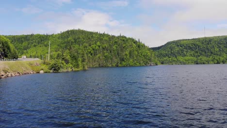 drone footage flying quickly out over the middle of a lake with forest-covered mountains in the background, a road on the left during the day