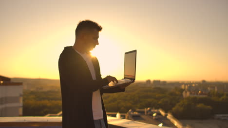 the camera follows a man walking across a rooftop at sunset with a laptop and a bottle. the freelancer opens the laptop and types on the keyboard. slow motion hacker walks on the roof with a laptop.