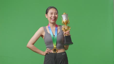 asian woman with a gold medal and trophy smiling to camera with one arm akimbo posing as the first winner on green screen background in the studio