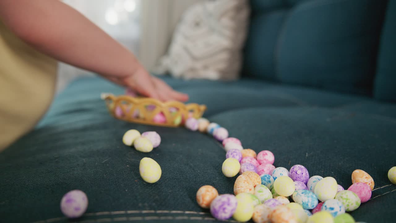 Child's hand pushes a basket of colorful candies across a couch. The basket tips, spilling candies onto the fabric. The child's hand moves gently, showing care and curiosity