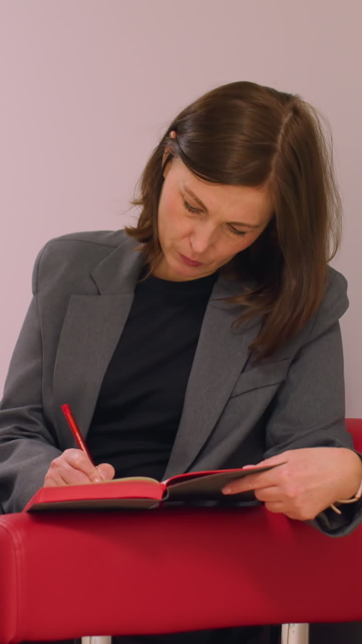 Businesswoman sitting on red chair in office hallway, writing in notebook, focused on task, preparing for meeting or planning, professional and calm environment, office decor in background