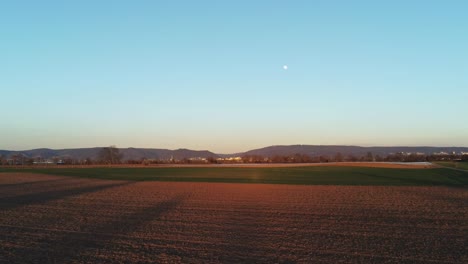 Alejarse-Del-Paisaje-Sobre-El-Campo-Agrícola-Durante-La-Puesta-De-Sol-Con-Luna-Llena-En-El-Cielo-En-Alemania,-Europa