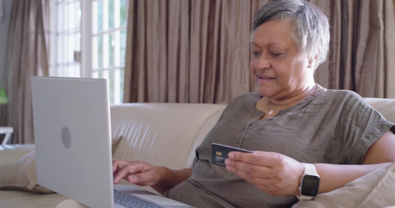 Anziana donna afroamericana che paga una bolletta a casa, tenendo una carta e digitando sul laptop, con lo smartwatch.