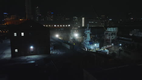 Forwards-fly-above-docks-at-waterfront.-Glowing-lights-shining-on-boats.-Night-city-in-background.-Manhattan,-New-York-City,-USA