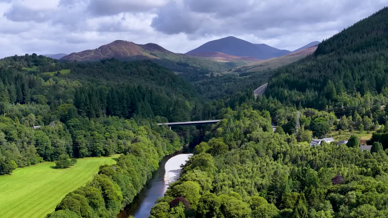 Drone glides over lush green valley, river, and bridge beneath dramatic clouds in Scottish Highlands