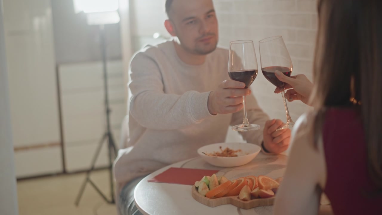 Uomo seduto solleva un bicchiere di vino verso la sua compagna dall'altra parte di un piccolo tavolo rotondo durante un pasto rilassato al chiuso, con antipasti impiattati e pane sul tavolo sotto una luce calda e soffusa e un'atmosfera accogliente.