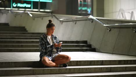 young woman using phone on city stairs at night