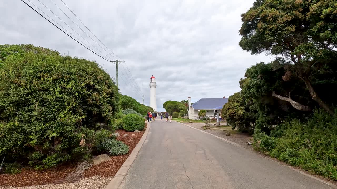 A serene walk along a tree-lined path towards Aireys Inlet Lighthouse under overcast skies, capturing natural beauty and tranquility