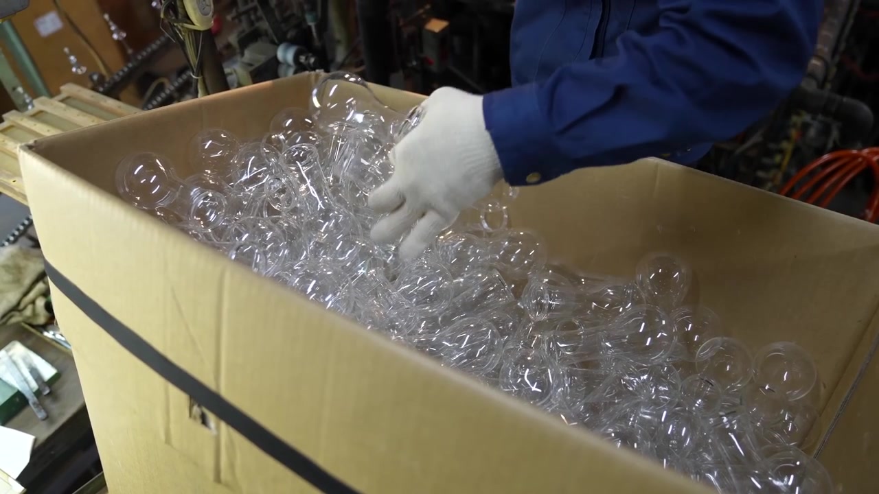 Operaio che smista involucri di lampadine in vetro soffiato all'interno di una grande scatola di cartone.