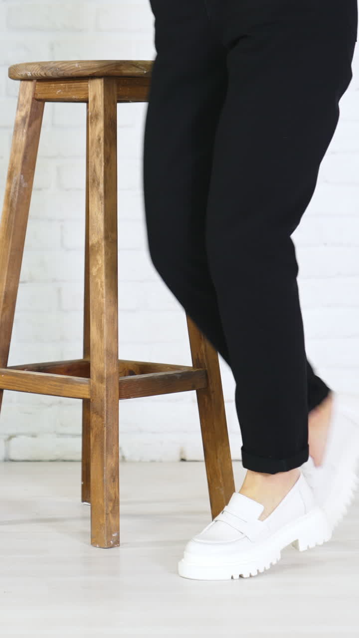 Model in black jeans demonstrating white massive shoes. Lady turns around to show the fashionable footwear. Wooden tabouret at backdrop. Vertical video