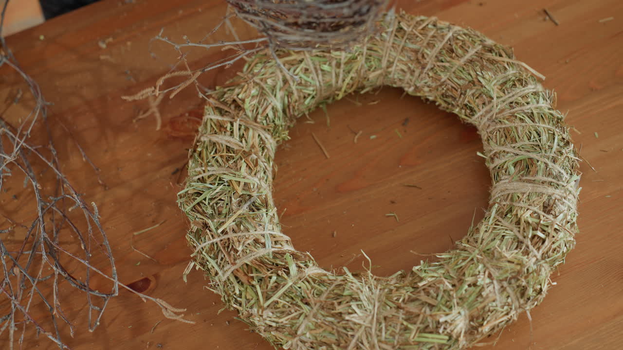 Female florist arranging natural twigs on straw wreath base, creating rustic handmade decoration on wooden table with moss, wood slices, and tools, capturing detailed artisan process of design