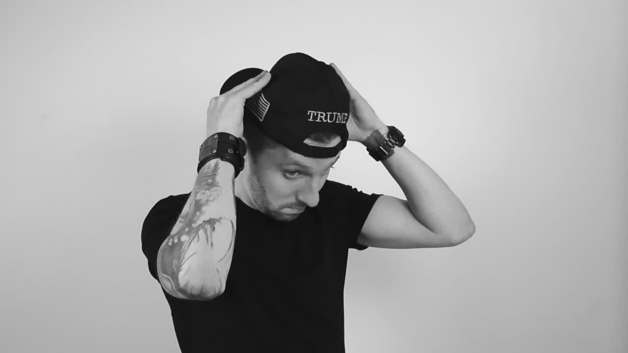 Grayscale footage of a man with tattoos putting on a dark black MAGA or Make America Great Again hat in reverse that has TRUMP written on it and start to nod while being happy Patriot Donald Trump fan
