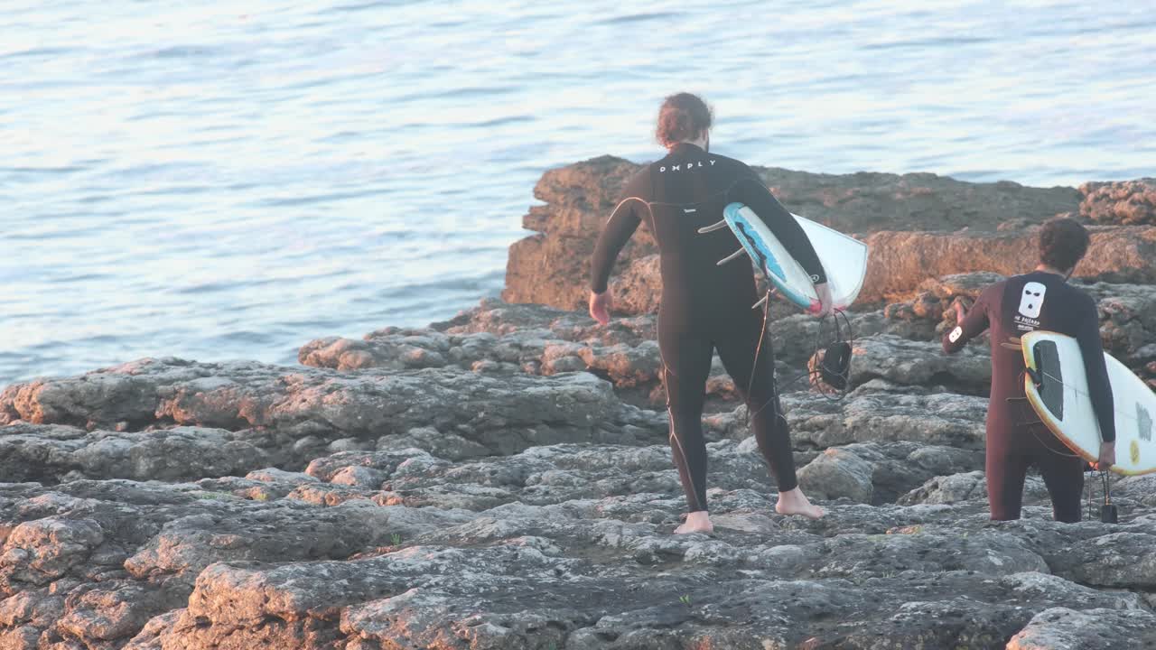 pasarela aérea con drones de dos surfistas esperando en un acantilado rocoso para saltar y surfear con olas encaladas en el lugar de surf de estoril