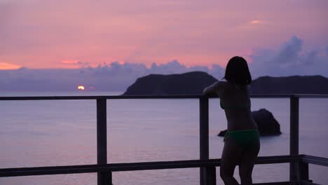 スローモーションを背景に島々が浮かぶ海に沈む夕日を眺めるビキニの女の子のシルエット