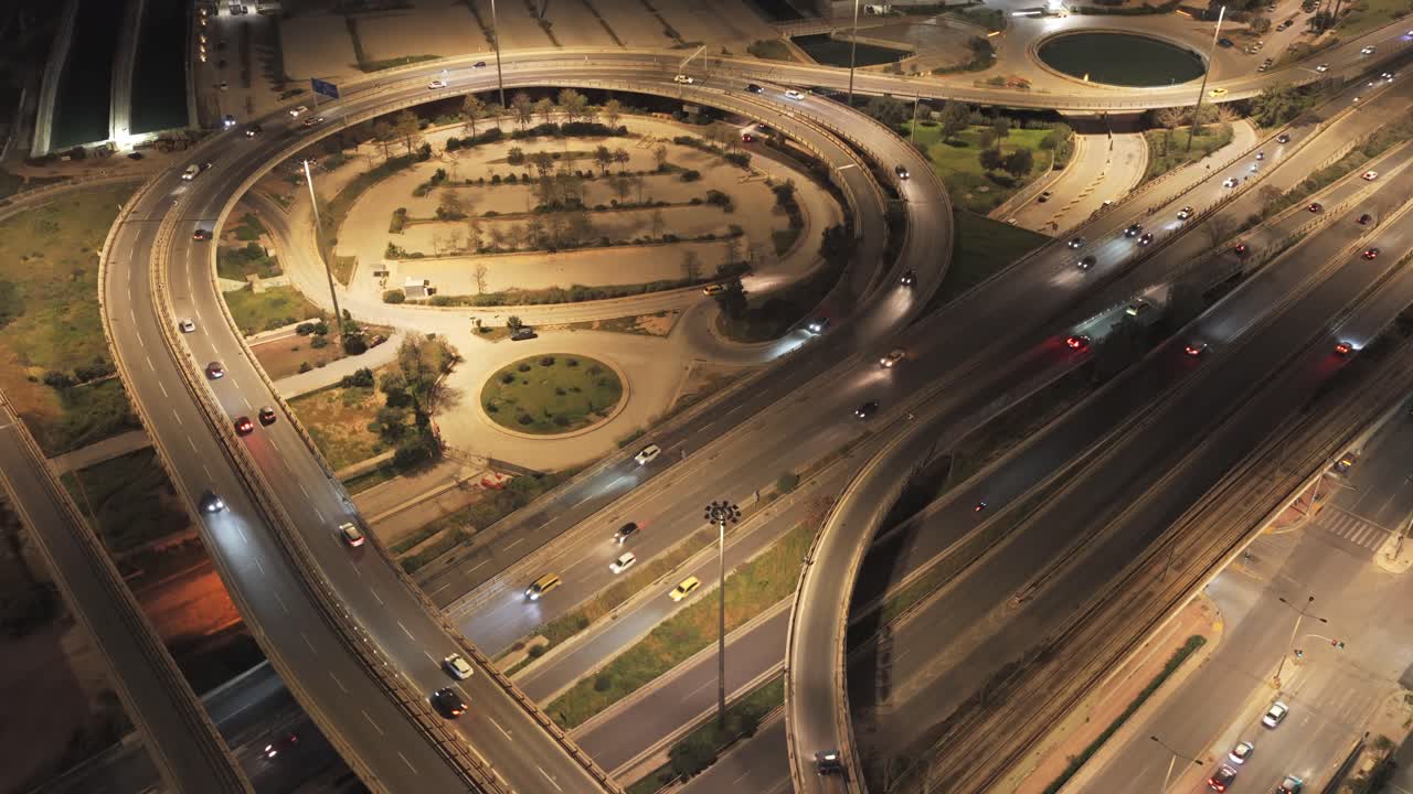 Aerial view of a bustling multi-level highway interchange with heavy traffic flow at night, showing illuminated roads, overpasses, and intricate road networks creating a dynamic urban landscape