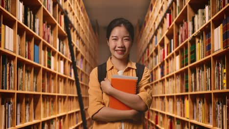 asian woman student with a backpack holding some books and smiling to camera while standing in the library